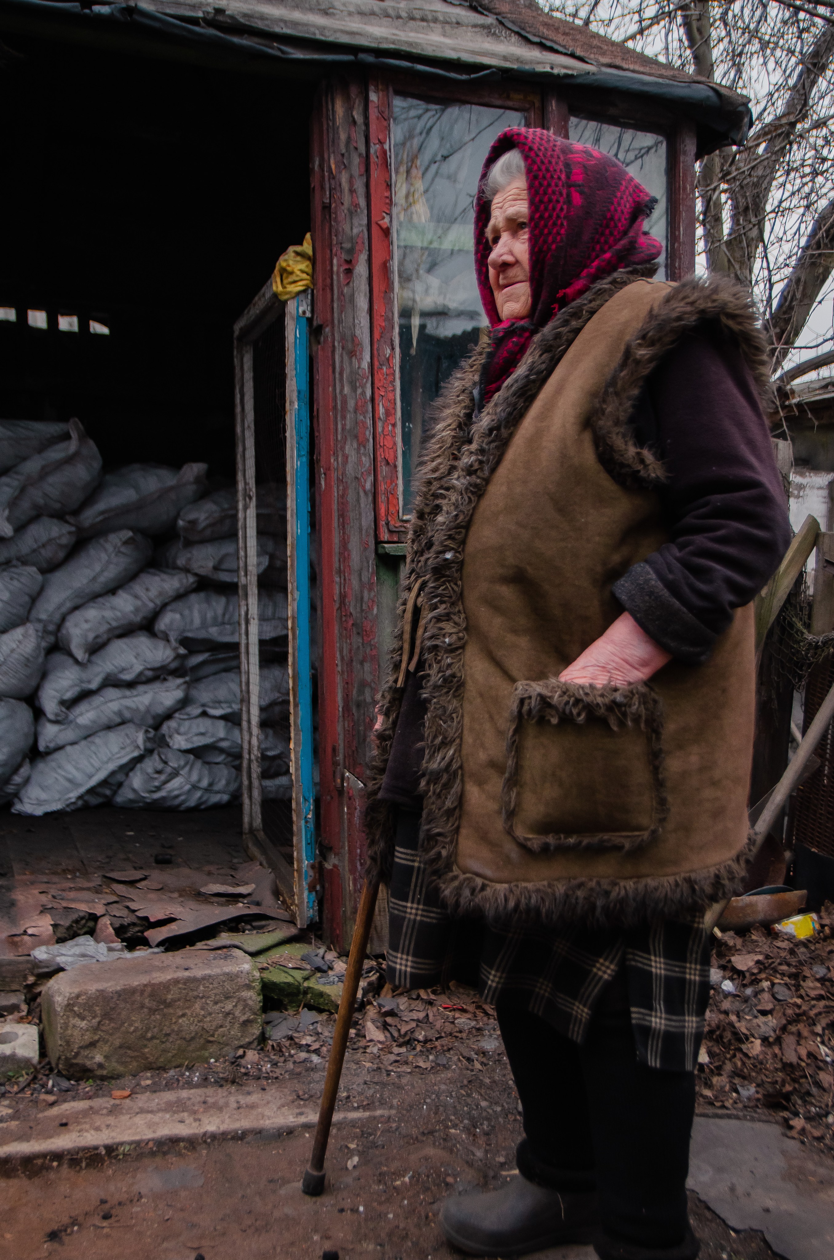 Пожилая женщина в коричневой меховой жилетке и платке стоит с тростью возле сарая с мешками.